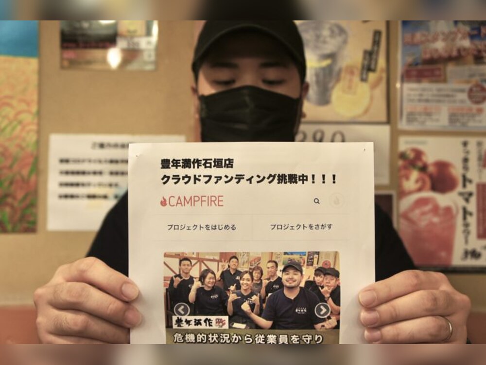 豊年満作 石垣店のオーナー「黒島」さんがクラウドファンディングのチラシを持って宣伝している様子